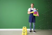 Male janitor with green cleaning supplies near wet floor sign